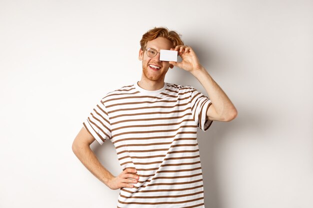 Concept d'achats et de finances. Heureux jeune homme aux cheveux rouges et lunettes montrant une carte de crédit en plastique près du visage, souriant joyeusement à la caméra, debout sur fond blanc.