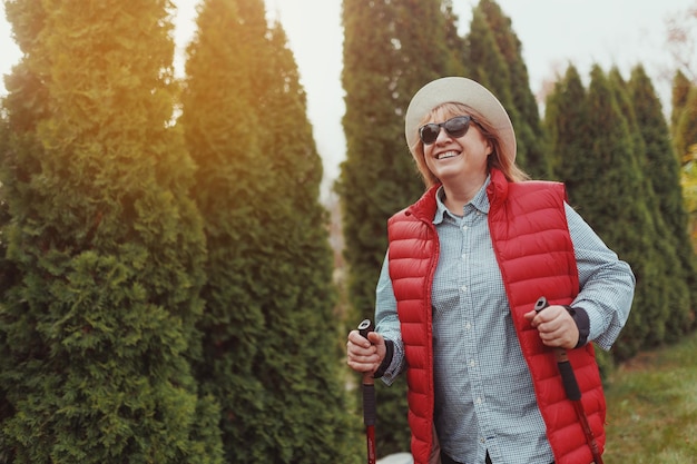 Concept d'activités de plein air personnes et vacances Jolie femme d'âge moyen aux cheveux courts en vêtements de sport randonnée en forêt à l'aide de bâtons pour la marche nordique faisant de l'exercice aérobique en profitant de la nature