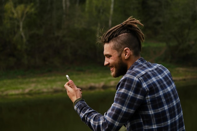 Concept d'affaires et de conversations téléphoniques Jeune homme de race blanche avec des dreadlocks dans le parc au bord de la rivière et parlant sur un lien vidéo souriant Communiquer en ligne à l'aide d'un appareil mobile