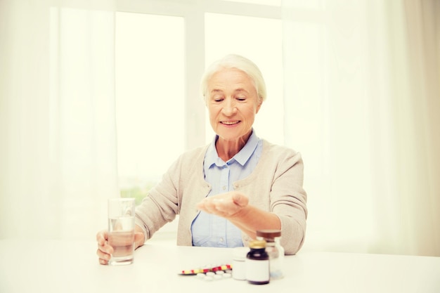 concept d'âge, de médecine, de soins de santé et de personnes - femme âgée heureuse avec des pilules et un verre d'eau à la maison