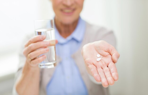 concept d'âge, de médecine, de soins de santé et de personnes - gros plan d'une femme âgée heureuse avec des pilules et un verre d'eau à la maison