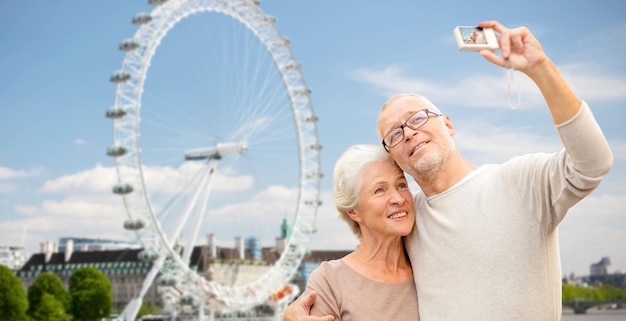 concept d'âge, de tourisme, de voyage, de technologie et de personnes - couple de personnes âgées avec caméra prenant selfie sur fond de roue de ferry de londres