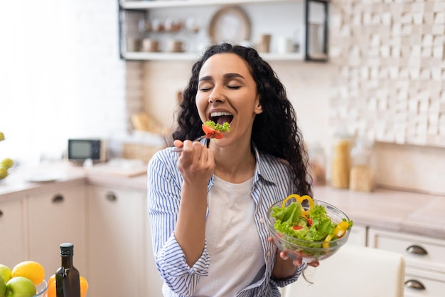 Concept d'alimentation saine femme excitée appréciant une salade de légumes frais en train de dîner dans la cuisine à la maison