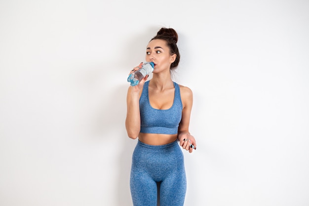 Le concept d'une alimentation saine. Une jeune femme blanche en tenue de sport, avec une silhouette tonique, buvant de l'eau d'une bouteille. Fond blanc. Espace de copie.