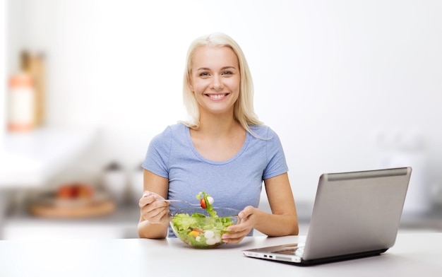 concept d'alimentation saine, de régime et de personnes - jeune femme souriante avec ordinateur portable mangeant une salade de légumes sur fond de cuisine