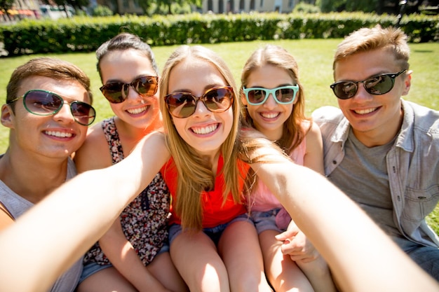 concept d'amitié, de loisirs, d'été, de technologie et de personnes - groupe d'amis souriants faisant du selfie avec un smartphone ou une tablette pc dans le parc