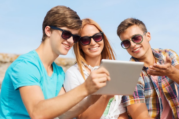 Concept d'amitié, de loisirs, d'été, de technologie et de personnes - groupe d'amis souriants avec un ordinateur tablette assis à l'extérieur