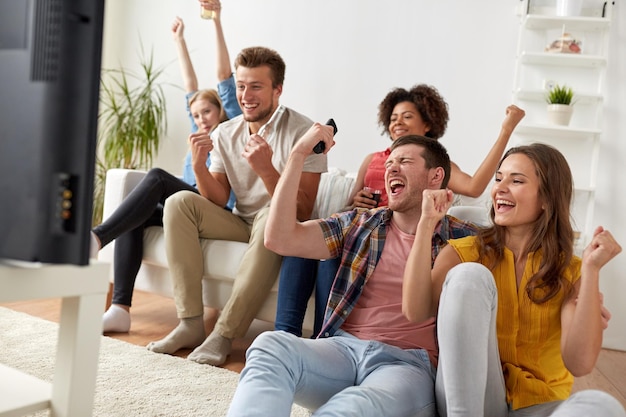 concept d'amitié, de loisirs, de personnes et de divertissement - amis heureux avec télécommande et boissons en regardant la télévision à la maison
