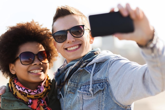 concept d'amitié, de relations, de tourisme, de voyage et de personnes - amis adolescents heureux ou couple à lunettes de soleil avec smartphone prenant selfie à l'extérieur