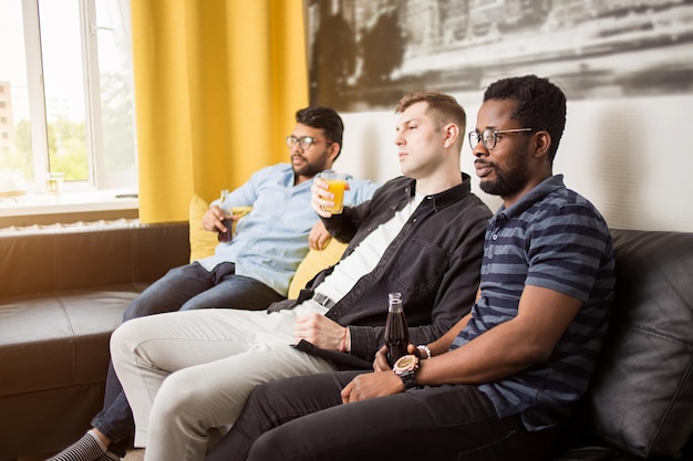 Concept d'amitié, de sport et de divertissement. Vue latérale de trois amis regardant la télévision en buvant des boissons gazeuses et du jus assis sur un canapé à la maison avec un intérieur moderne.