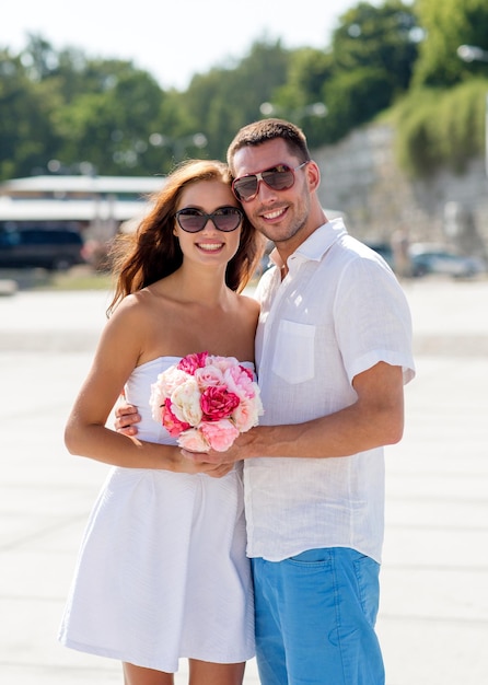 concept d'amour, de mariage, d'été, de rencontres et de personnes - couple souriant portant des lunettes de soleil avec bouquet de fleurs étreignant dans la ville