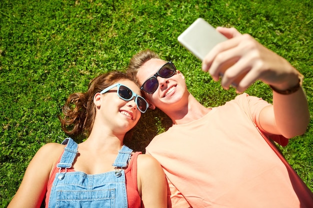 concept d'amour et de personnes - couple d'adolescents heureux en lunettes de soleil allongé sur l'herbe et prenant selfie sur smartphone en été