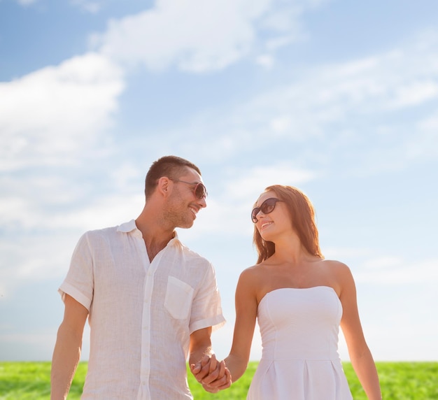 concept d'amour, de personnes, d'été et de relations - couple souriant portant des lunettes de soleil marchant à l'extérieur sur fond de ciel bleu et d'herbe