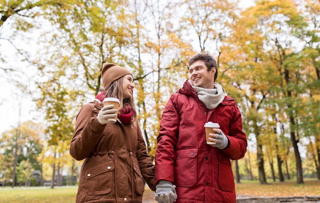 concept d'amour, de relations, de saison et de personnes - jeune couple heureux avec des tasses de café marchant dans le parc d'automne