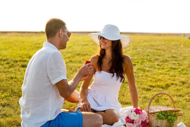 concept d'amour, de rencontres, de personnes et de vacances - jeune homme souriant montrant une petite boîte cadeau rouge à sa petite amie en pique-nique