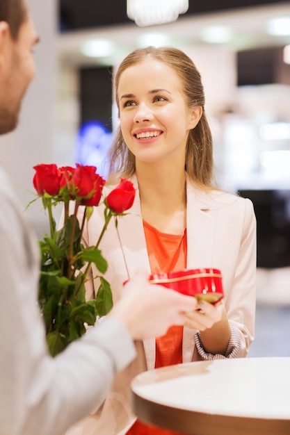 concept d'amour, de romance, de saint valentin, de couple et de personnes - jeune homme heureux avec des fleurs rouges donnant un cadeau à une femme souriante au café du centre commercial