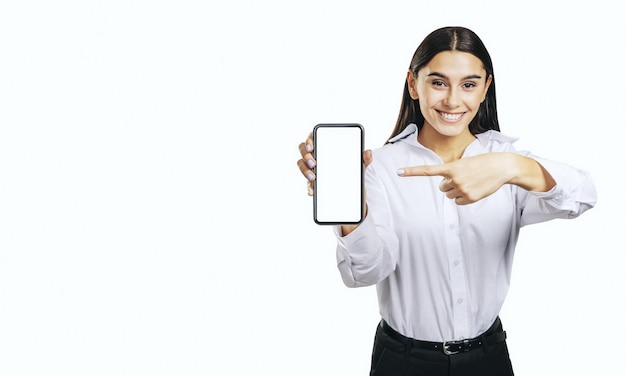 Concept d'application mobile avec une fille heureuse en chemise blanche montrant un smartphone moderne avec un écran blanc vierge sur fond clair abstrait avec place pour votre logo ou maquette de texte