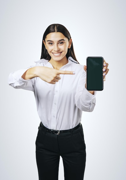 Concept d'application mobile avec une fille heureuse en chemise blanche montrant un téléphone portable moderne avec un écran blanc sur une maquette abstraite de fond clair
