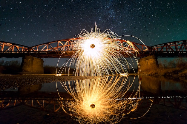 Concept d'art de peinture légère. Filature de laine d'acier dans un cercle abstrait, feux d'artifice de brillants jaune vif scintille sur un long pont reflété dans l'eau de la rivière sur le ciel étoilé de la nuit bleue