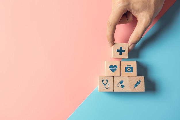 Concept D'assurance Maladie, Main De L'homme Organisant L'empilement De Bois Cube Avec Icône De Santé Médical Sur Fond Bleu Et Rose Pastel, Espace Copie.