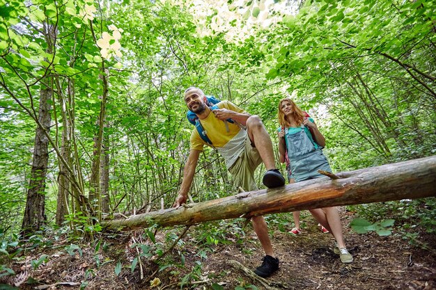 concept d'aventure, de voyage, de tourisme, de randonnée et de personnes - groupe d'amis souriants marchant avec des sacs à dos et grimpant sur un tronc d'arbre tombé dans les bois