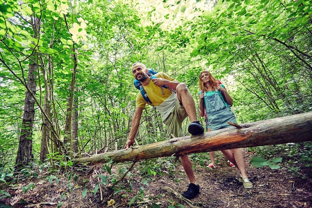 concept d'aventure, de voyage, de tourisme, de randonnée et de personnes - groupe d'amis souriants marchant avec des sacs à dos et grimpant sur un tronc d'arbre tombé dans les bois