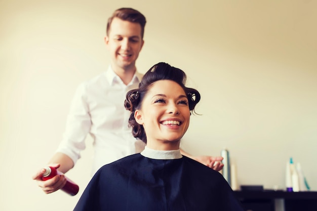 concept de beauté, de coiffure et de personnes - jeune femme heureuse avec un coiffeur avec une laque pour cheveux fixant la coiffure au salon