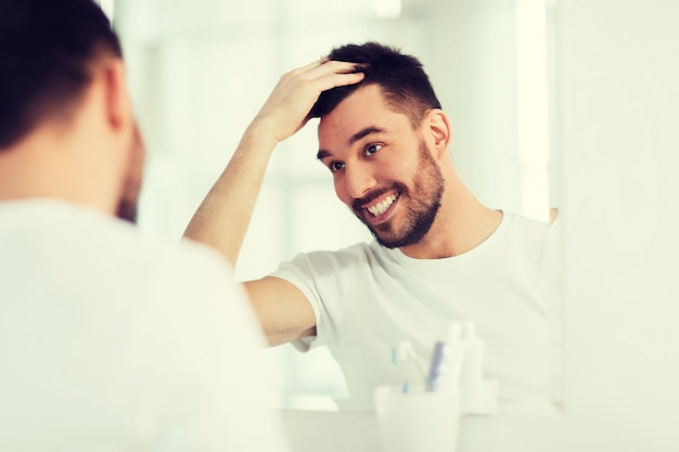 Photo concept de beauté, d'hygiène, de coiffure et de personnes - jeune homme souriant à la recherche de miroir et de coiffure à la salle de bain à la maison