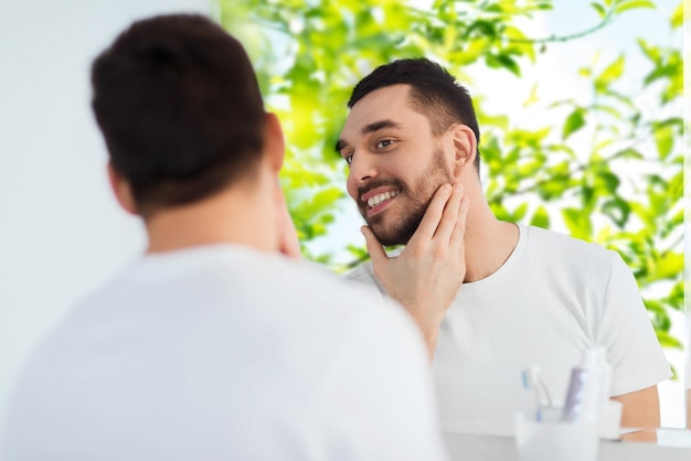 concept de beauté, d'hygiène et de personnes - jeune homme souriant regardant un miroir dans la salle de bain à la maison sur fond naturel vert