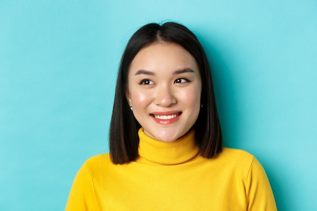 Concept de beauté et de maquillage. Fille asiatique de rêve avec une peau parfaitement saine, regardant à gauche et souriante heureuse, debout sur fond bleu