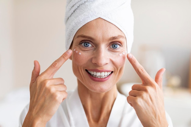 Concept de beauté mature Charmante femme d'âge moyen appliquant de la crème pour le visage sous ses yeux et souriant à la caméra