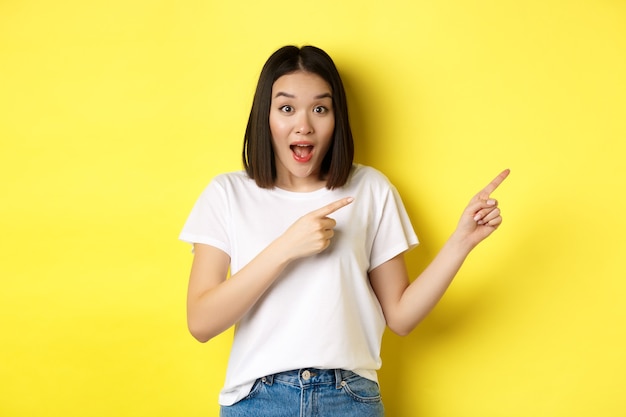 Concept de beauté et de mode. Belle femme asiatique en t-shirt blanc pointant les doigts à droite, démontrer le logo debout sur fond jaune.