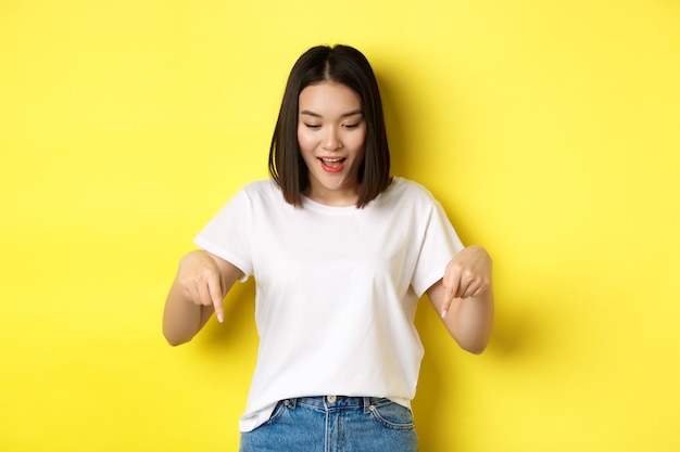 Concept de beauté et de mode. Belle femme asiatique en t-shirt blanc pointant les doigts vers le bas, démontrer le logo debout sur fond jaune.