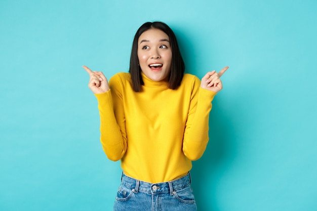 Concept de beauté et de mode. Excité fille asiatique en pull jaune faisant son choix, pointant du doigt sur le côté et choisissant entre deux options, debout sur fond bleu.