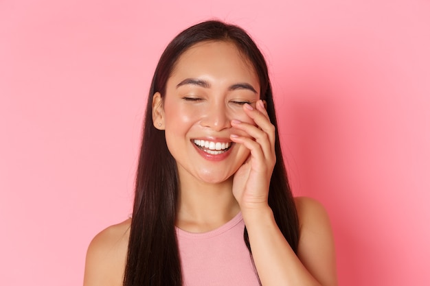 Concept de beauté, mode et style de vie. Gros plan d'une belle fille asiatique insouciante touchant le visage et riant joyeusement les yeux fermés, debout sur le mur rose joyeux, promotion de produit de soin de la peau.