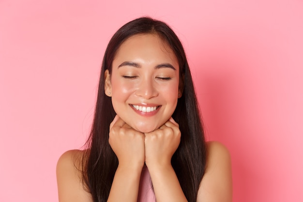 Concept de beauté, mode et style de vie. Gros plan d'une fille asiatique rêveuse et romantique, séduisante, rêvasser, visage appuyé sur les mains et fermer les yeux avec un sourire blanc heureux, mur rose.