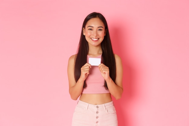 Concept de beauté, mode et style de vie. Portrait de belle fille asiatique à la mode montrant sa carte de crédit bancaire et souriant, recommander un compte de démarrage ou consulter la nouvelle fonctionnalité e-banking, mur rose