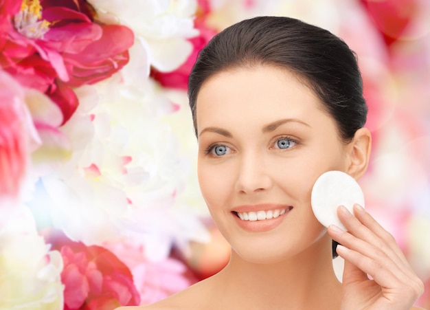 concept de beauté, de personnes et de santé - belle femme souriante nettoyant la peau du visage avec un tampon de coton sur fond floral rose