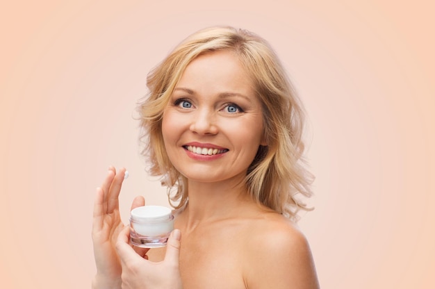 concept de beauté, de personnes, de soins de la peau et de cosmétiques - femme heureuse appliquant de la crème sur son visage sur fond beige