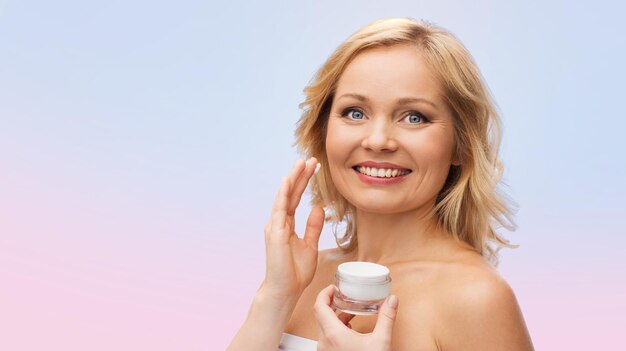 concept de beauté, de personnes, de soins de la peau et de cosmétiques - femme heureuse appliquant de la crème sur son visage sur fond dégradé de quartz rose et de sérénité