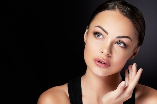 Photo concept de beauté, portrait de la tête et des épaules du beau modèle avec maquillage nu à la recherche de côté et vers le haut, toucher le visage, gros plan. femme brune en studio avec fond noir