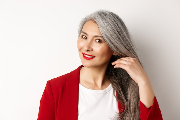 Concept de beauté et de soins capillaires. Gros plan d'une femme âgée asiatique élégante montrant des cheveux gris brillants et sains, souriant et regardant de côté, fond blanc