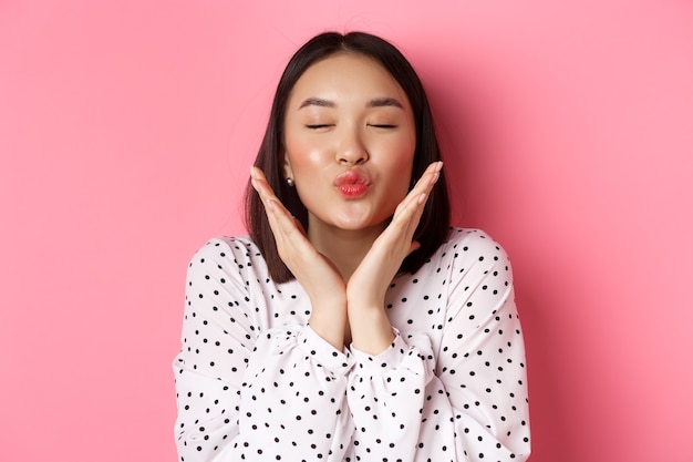 Concept de beauté et de soins de la peau. Gros plan sur une belle fille asiatique ferme les yeux, des lèvres plissées pour les baisers, montrant un joli visage parfait, debout sur fond rose