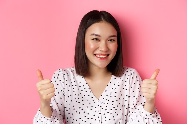 Concept de beauté et de style de vie. Gros plan d'une jolie femme asiatique montrant son soutien, faisant un geste de pouce levé et souriant, comme et vous féliciter, debout sur fond rose