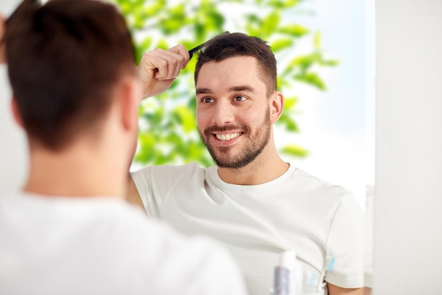 concept de beauté, de toilettage et de personnes - jeune homme souriant regardant au miroir et se brossant les cheveux avec un peigne à la maison salle de bain sur fond naturel vert