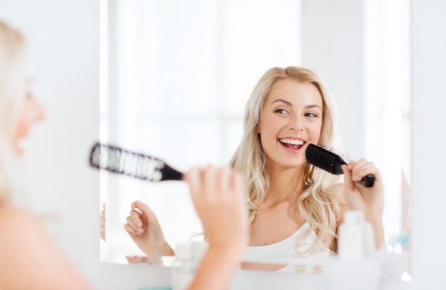 concept de beauté, de toilettage, de soins capillaires et de personnes - jeune femme souriante regardant au miroir et chantant à la brosse à cheveux ou au peigne à la salle de bain à la maison
