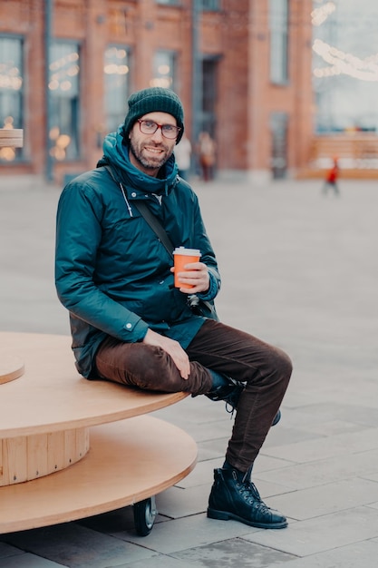 Concept de boissons et de style de vie Un homme européen insouciant et satisfait avec des poils profite d'une journée ensoleillée et des sourires de boissons chaudes s'assoit joyeusement sur fond de rue Photo en plein air d'un beau hipster