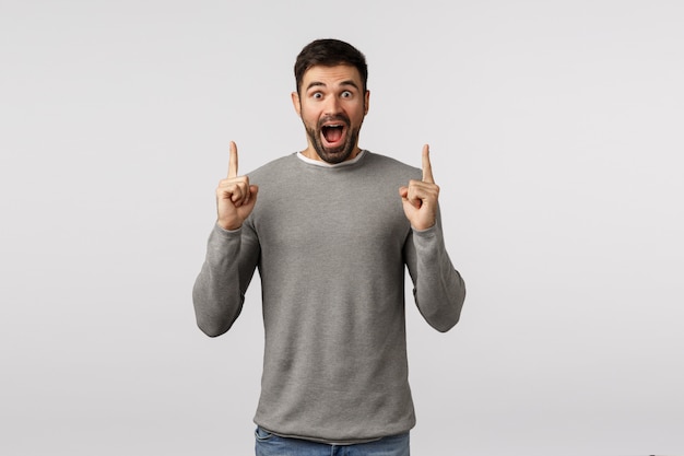 Concept de bonheur, de joie et de surprise. Homme barbu étonné, sans voix et émerveillé en pull gris, voir quelque chose d'incroyable et incroyable, pointant les doigts vers le haut