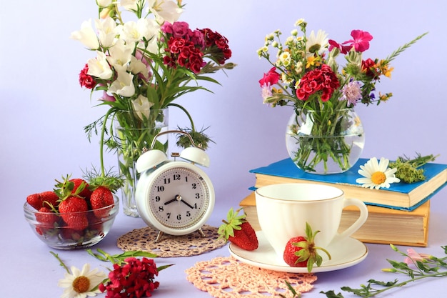 Le concept de bonjour et de bonne humeur Livres une tasse de thé fleurs sauvages dans des vases fraises dans et autour d'une tasse en verre une horloge sur fond clair