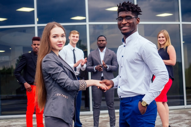 Concept de bonne affaire. Groupe multinational de jeunes gens d'affaires salutations et poignée de main avec des collègues en arrière-plan. Belle femme d'affaires souriante avec un partenaire afro-américain se serrant la main